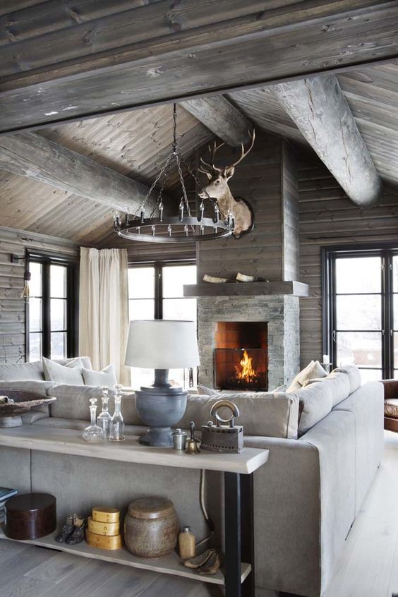 a neutral chalet living room with stained wood and wooden beams, a stone fireplace, a neutral sectional, a console table and a metal chandelier