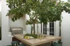 a quirky outdoor dining area with a stone table and a livign tree inside it plus dark green stools and a grill next to it