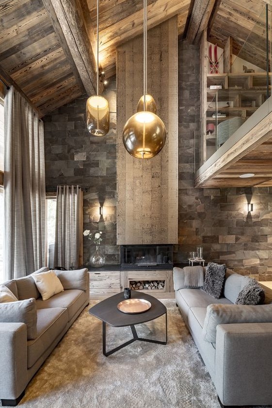 a refined chalet living room with a stone wall, a fireplace, a wooden ceiling, grey sofas and hanging glass lamps