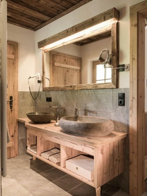 a rustic chalet bathroom done with blonde wood and rough stone sinks, with a large lit up mirror and a vanity on legs