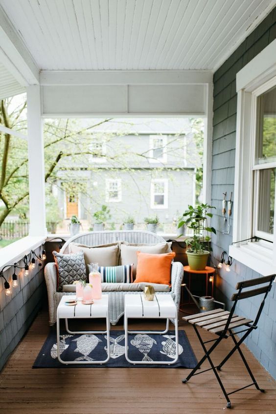 a simple summer porch with a white wicker loveseat, white stools, a wooden chair and some lights around