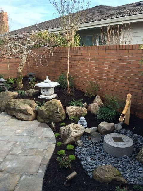 un pequeño jardín japonés con grandes rocas, una fuente de bambú, guijarros, un cuenco de piedra y árboles, además de una gran linterna de piedra