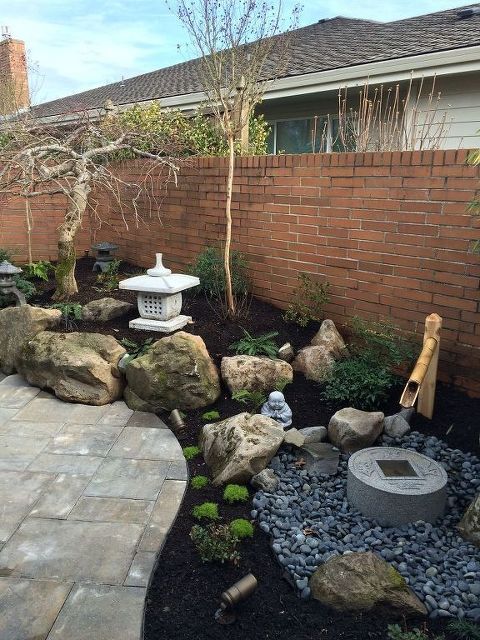 a small Japanese garden with large rocks, a bamboo fountain, pebbles, a stone bowl and trees plus a large stone lantern