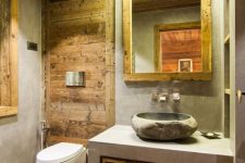 a small chalet bathroom clad with cocnrete and with lots of wood, with a mirror in a wooden frame and a built-in vanity