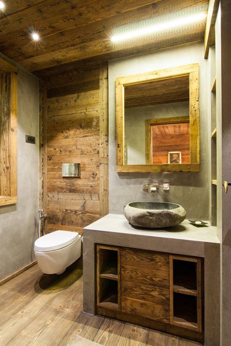 a small chalet bathroom clad with cocnrete and with lots of wood, with a mirror in a wooden frame and a built-in vanity