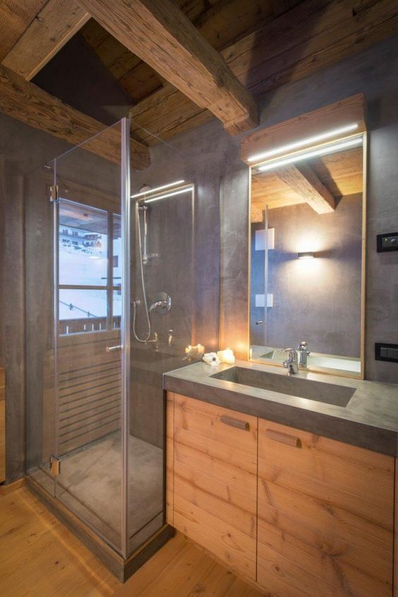 a small minimalist chalet bathroom done with blonde wood and concrete, with textural wooden beams on the ceiling and a lit up mirror