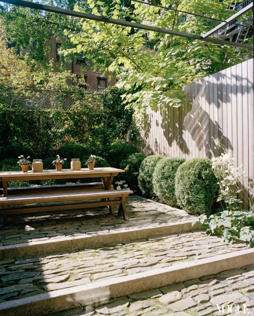 a townhouse garden with stoen steps, greenery and boxwood growing and a simple wooden dining set with benches