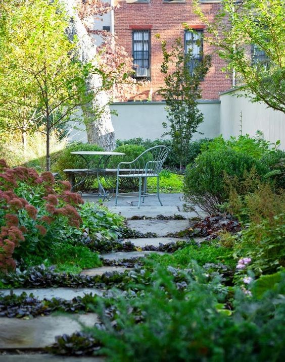 a townhouse garden with trees and lush greenery growin even in between the stone steps and metal furniture