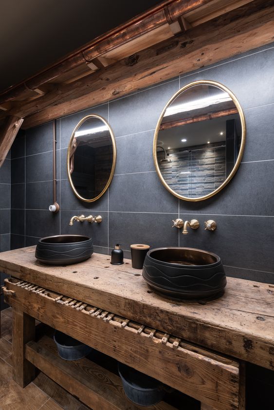 an exquisite chalet bathroom with grey tiles, round mirrors, round black bowl sinks, brass fixtures and a wooden vanity