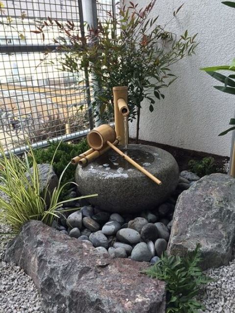 rocks, pebbles, a traditional stone and bamboo fountain, greenery in between the rocks compose a small and lovely front yard