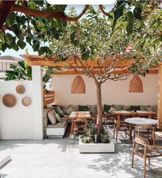 a Mediterranean dining space with a roof with greenery, a corner sofa with grey and green pillows, wicker pendant lamps and a tree in a planter