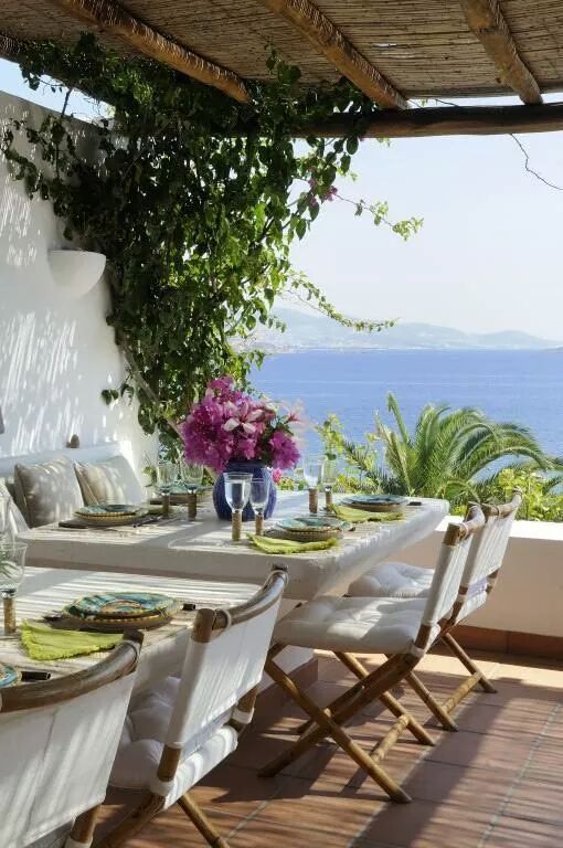 a Mediterranean dining space with a view, with a built-in sofa, white tables and folding chairs with white upholstery, blooms and greenery