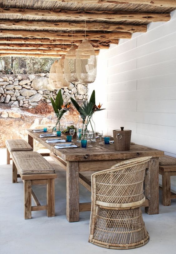 a Mediterranean dining spot with a roof with pendant lamps, stained dinign set with benches, a wicker chair and blue glasses