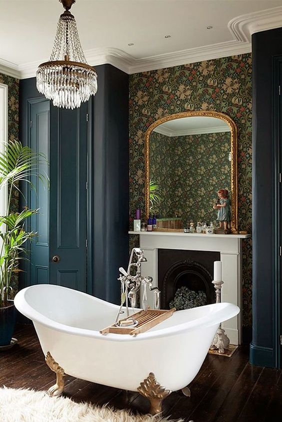 a Victorian style bathroom with moody wallpaper walls, a non-working fireplace, a clawfoot bathtub, navy wardrobes, a crystal chandelier and plants