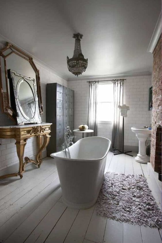 a beautiful vintage bathroom with a brick fireplace, a gold carved console table and mirrors, a free-standing bathtub, a crystal chandelier