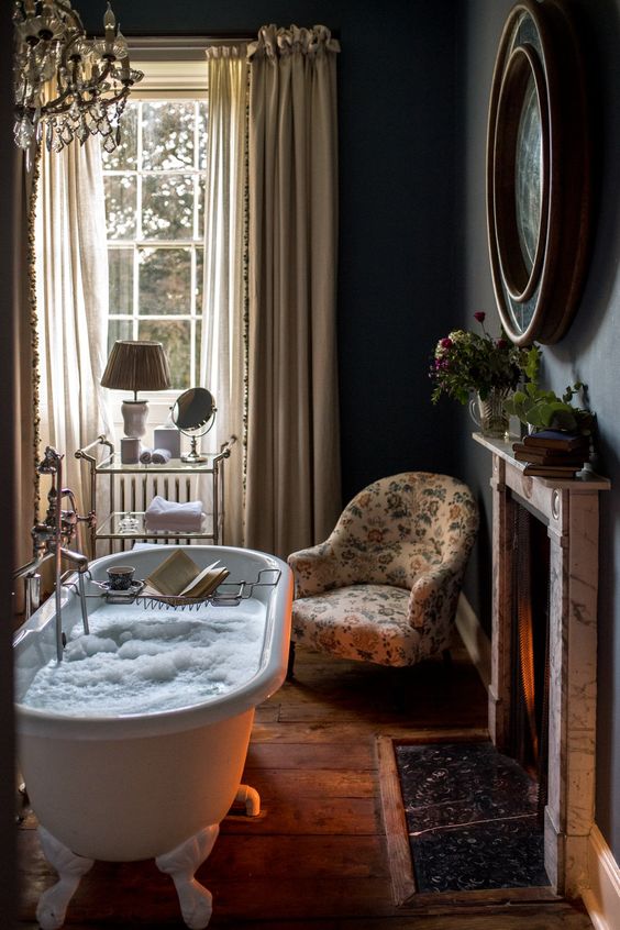 a beautiful vintage bathroom with black walls, a fireplace, a clawfoot bathtub, neutral curtains, a floral chair and some greenery and blooms