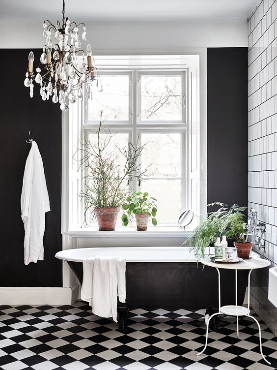 a black and white vintage bathroom with white square tiles, a black and white floor, a black statement wall, a crystal chandelier and lots of greenery