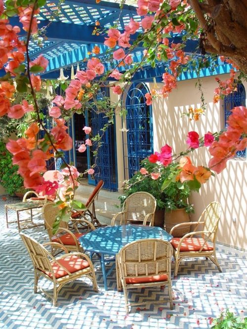 a bright Mediterranean dining space with a blue round table, wicker chairs with coral cushions, bright blooms over the space and blue window frames