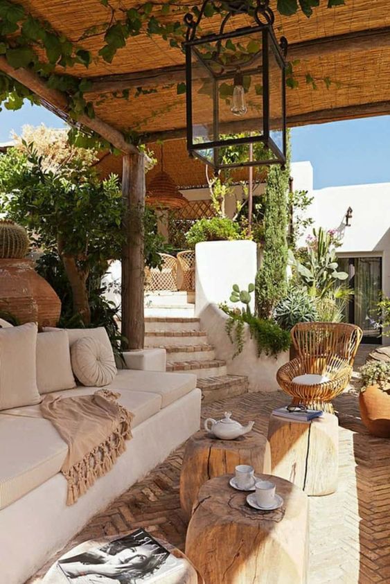 a chic Mediterranean patio done in neutrals, with a roof, greenery, a creamy sofa and pillows, stumps and a wicker chair, pendant lamps