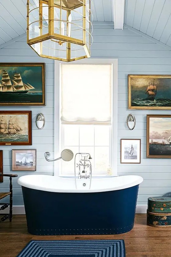 a chic and fancy bathroom with light blue planked walls, a navy tub and a fantastic marine gallery wall plus a glam gold lamp