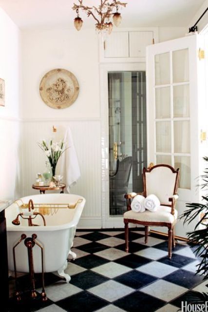 a chic vintage bathroom with a checked black and white floor, a white clawfoot bathtub, a creamy chair, some plants and a chic chandelier