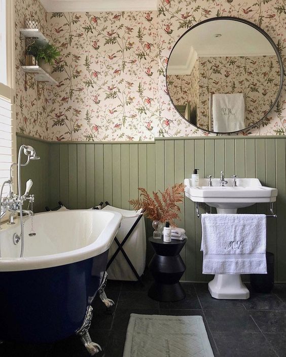 a cozy English country bathroom with floral wallpaper, green panels, a navy clawfoot bathtub, a free-standing sink and side tables