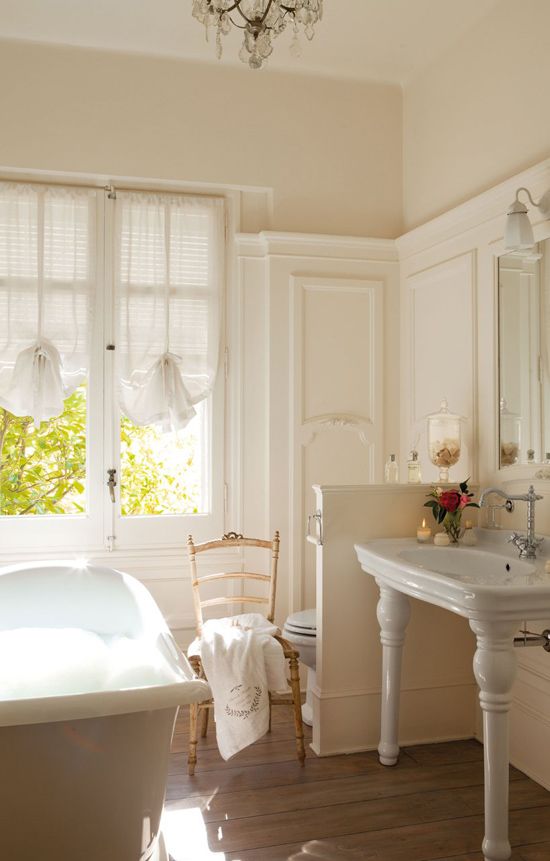 a delicate neutral bathroom with creamy paneling, a free-standing sink, a light-stained chair, a bathtub and a crystal chandelier