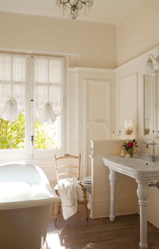 a delicate neutral bathroom with creamy paneling, a free-standing sink, a light-stained chair, a bathtub and a crystal chandelier