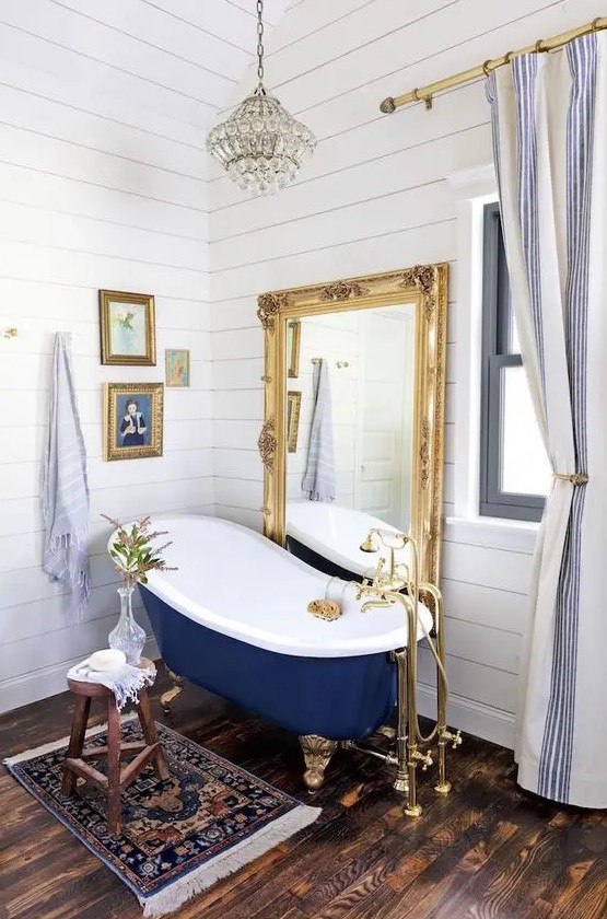 a fancy bathroom with white planked walls, a dark stained floor, a vintage navy bathtub, a floor mirror in a gilded frame, a mini gallery wall and a crystal pendant lamp