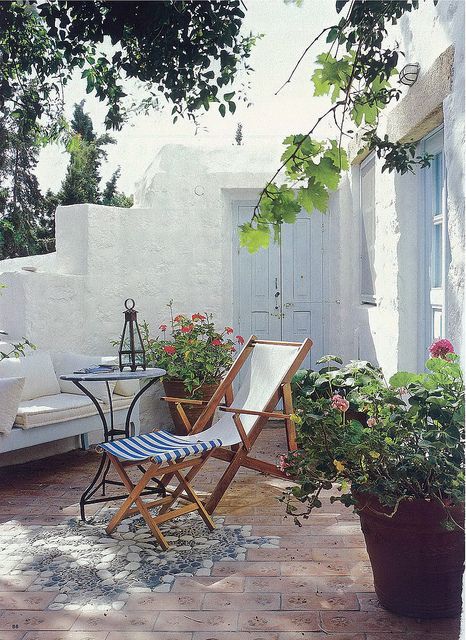 a little Mediterrranean outdoor nook with a sofa with white cushions and pillows, folding chairs, a metal table, bright blooms in planters