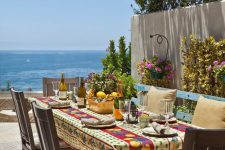 a lovely Mediterranean dining space with a view, a wooden table and chairs, a blue bench, a bright tablecloth and bright blooms