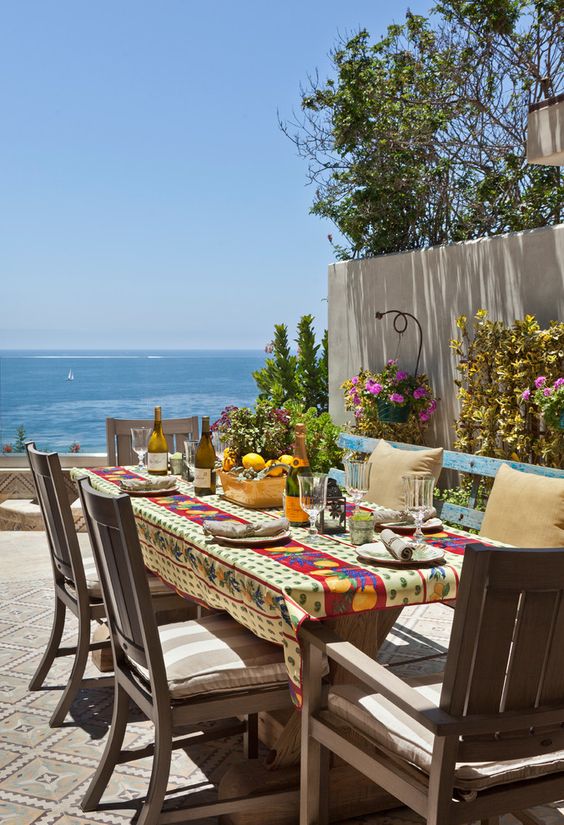 a lovely Mediterranean dining space with a view, a wooden table and chairs, a blue bench, a bright tablecloth and bright blooms