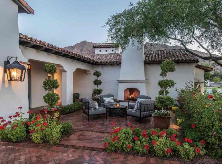 a lovely Mediterranean outdoor living room with greenery and blooms around, a fireplace, grey chairs, a black coffee table is welcoming