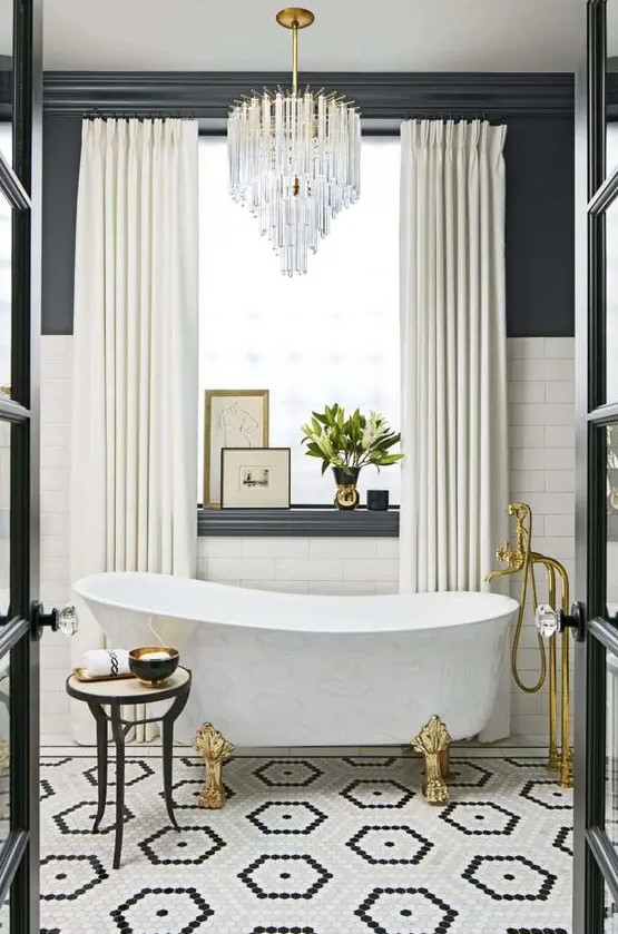 a luxorious bathroom with a black and white tile floor, white subway tiles and black walls, a chic tub, gold fixtures and a lovely chandelier