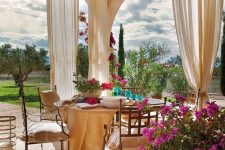 a neutral patio with neutral curtains, a round table and metal chairs, bright blooms and greenery and a view of the garden
