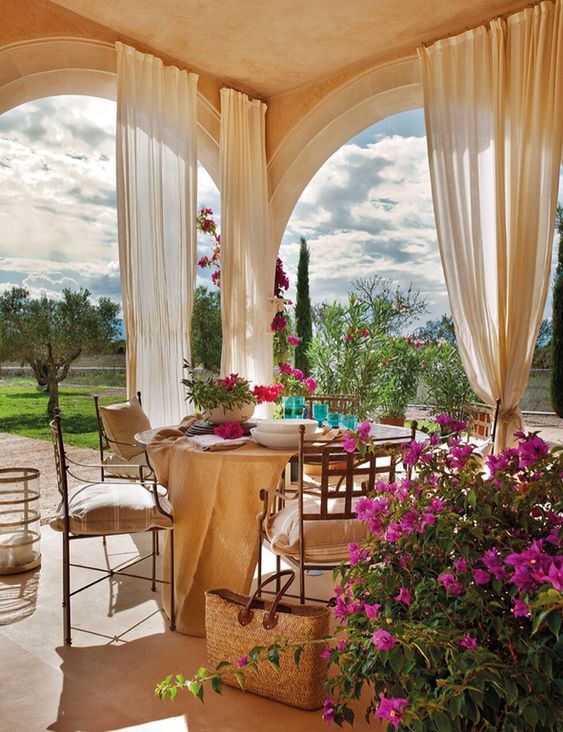 a neutral patio with neutral curtains, a round table and metal chairs, bright blooms and greenery and a view of the garden
