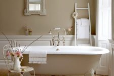 a neutral vintage bathroom with tan walls, white paneling, a tan clawfoot bathtub, a metal chair, a ladder and a mirror in an ornated frame