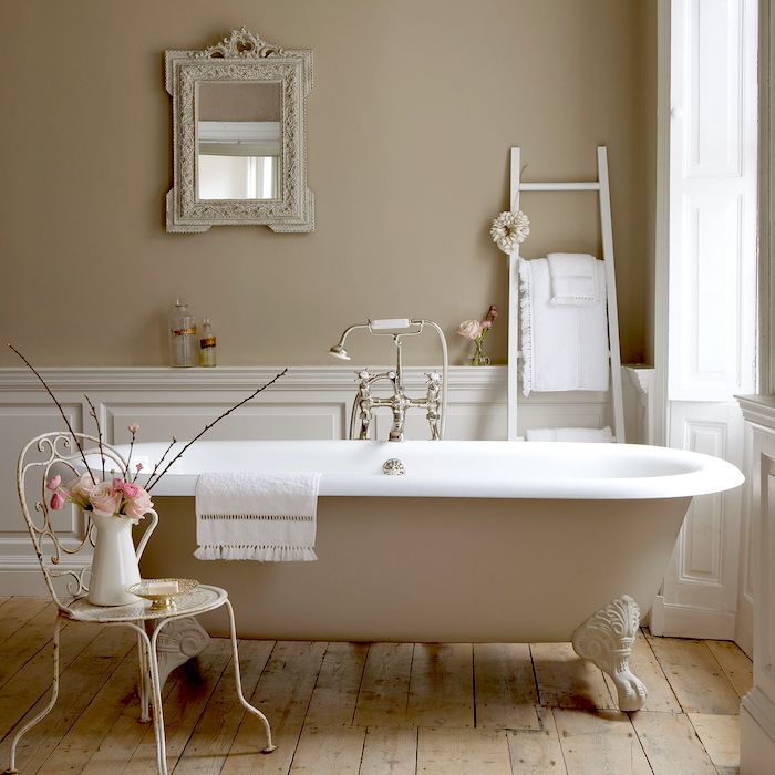 a neutral vintage bathroom with tan walls, white paneling, a tan clawfoot bathtub, a metal chair, a ladder and a mirror in an ornated frame