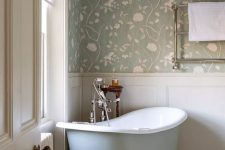 a pastel and neutral cottage bathroom with floral wallpaper, creamy paneling, a light blue clawfoot bathtub, a pendant lamp and a vintage stool