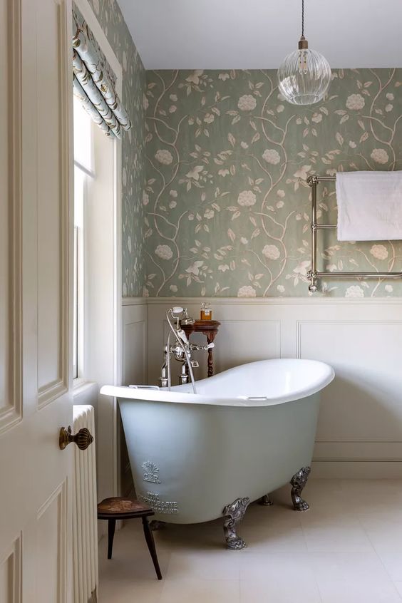 a pastel and neutral cottage bathroom with floral wallpaper, creamy paneling, a light blue clawfoot bathtub, a pendant lamp and a vintage stool