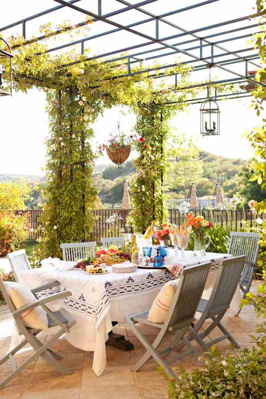 a pretty and livly Mediterranean terrace with a large table, grey chairs, greenery and blooms around and a lovely view