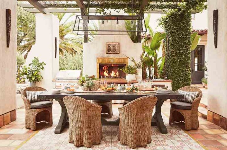 a refined Mediterranean outdoor dining space with greenery and potted plants, a dark-stained table and wicker chairs, a pendant lamp and a fireplace