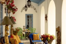 a refined Mediterranean patio done in bright colors, with a dining zone with a round table and green chairs, a glass table and bold blue wicker chairs, bright blooms and pendant lanterns