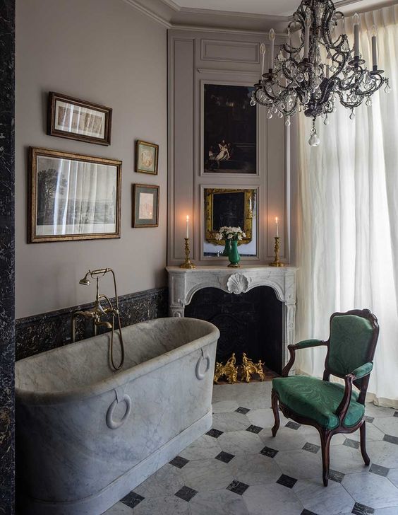 a refined vintage bathroom with a chic faux fireplace, a marble bathtub, a black chandelier, a gallery wall and a green chair