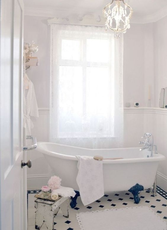 a subtle neutral bathroom with neutral walls, paneling, a clawfoot bathtub, a crystal chandelier, a black and white floor and neutral textiles