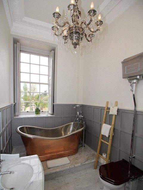 a vintage bathroom with grey paneling and shutters, a vintage metal bathtutb, a crystal chandelier, a fre-standing sink and a ladder for towels