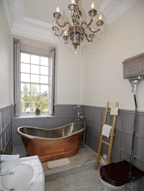 a vintage bathroom with grey paneling and shutters, a vintage metal bathtutb, a crystal chandelier, a fre-standing sink and a ladder for towels