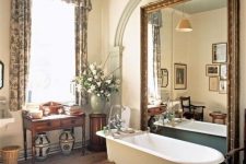 a vintage countryside bathroom done in neutrals, with an oversized mirror, floral curtains, vintage carved furniture, a clawfoot bathtub and pendant lamps