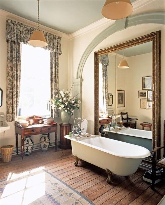 a vintage countryside bathroom done in neutrals, with an oversized mirror, floral curtains, vintage carved furniture, a clawfoot bathtub and pendant lamps