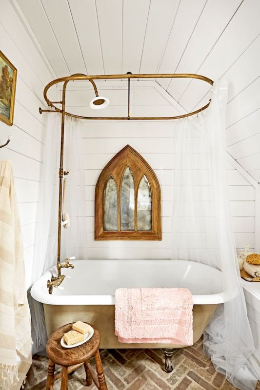 a vintage rustic bathroom with a brick floor, white shiplap on the walls, a tan clawfoot bathtub, a cnaopy and some wooden stools